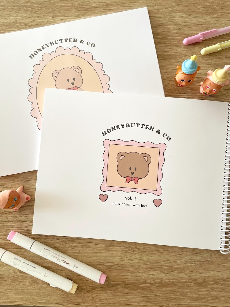 May include: White spiral-bound notebook and loose paper with a teddy bear illustration in a pink frame. The text "HONEYBUTTER & CO" and "vol. 1 hand drawn with love" are visible. Two markers and small figurines are also present.