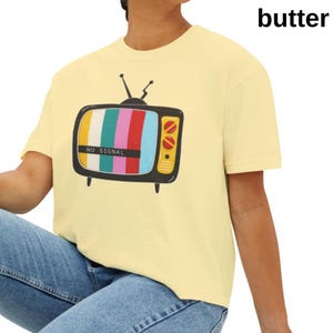 Könnte beinhalten: Hellgelbes T-Shirt mit einem Retro-Fernseher-Motiv. Der Fernsehbildschirm zeigt ein farbenfrohes Muster und die Worte "NO SIGNAL". Das T-Shirt ist kurzärmlig und hat eine lockere Passform.