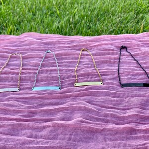 May include: Four necklaces with bar pendants are displayed on a pink cloth against a backdrop of green grass. The necklaces are gold, silver, and black. Each necklace has a chain and a rectangular bar pendant.