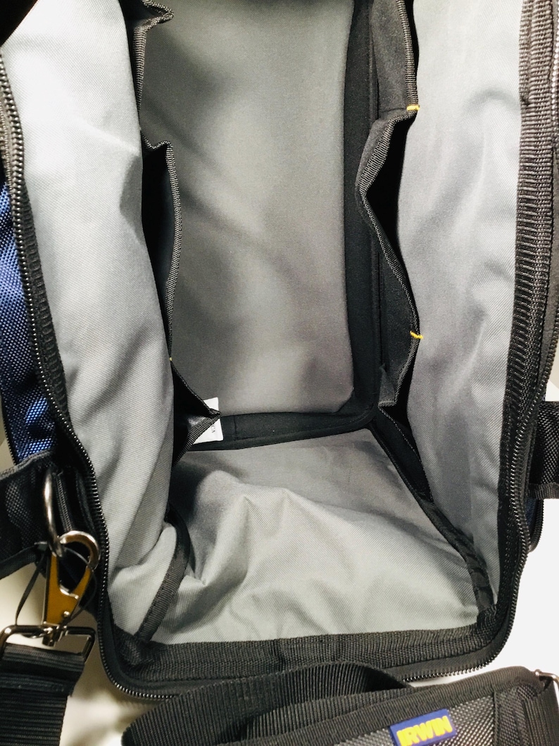 May include: Black and blue tool bag with multiple interior pockets and a gray lining. The bag has a black zipper, a shoulder strap, and metal hardware. The bag is designed for carrying tools and equipment.