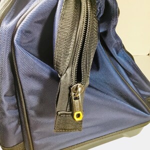 May include: Close-up of a navy blue rolling tool bag with black trim and zippers. The bag has a textured fabric and a yellow accent on the zipper pull. The bag appears to be new and unused.