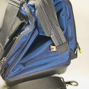May include: A blue and black nylon tool bag with a black shoulder strap and silver-tone metal hardware. The bag has multiple zippered compartments and a structured design. The bag is designed for carrying tools and equipment.