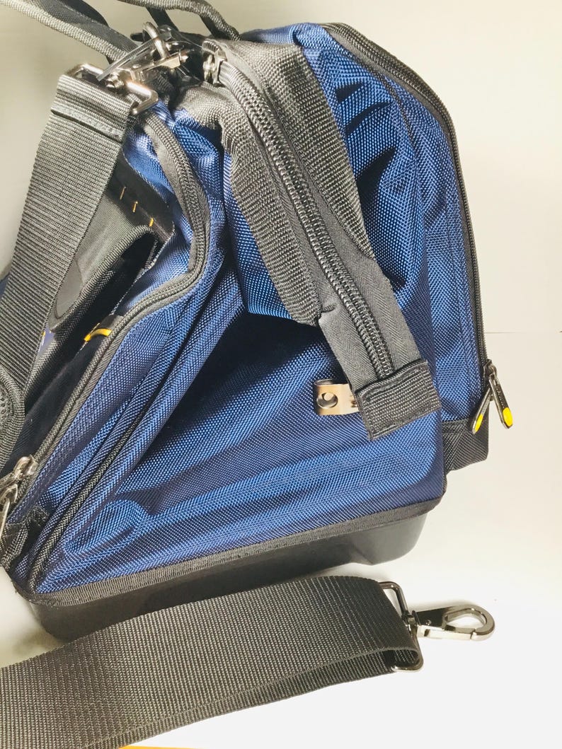 May include: A blue and black nylon tool bag with a black shoulder strap and silver-tone metal hardware. The bag has multiple zippered compartments and a structured design. The bag is designed for carrying tools and equipment.