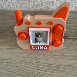 Könnte beinhalten: Ein pfirsich- und orangefarbenes 3D-gedrucktes Spielzeugradio mit einem zentralen quadratischen Rahmen, der ein Foto eines Kätzchens enthält. Das Gerät verfügt über eine Antenne, drei kleine Fischsymbole, zwei runde Knöpfe und ein weißes Namensschild mit dem Text LUNA.
