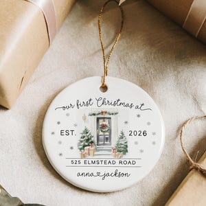 May include: A round white ceramic ornament featuring a watercolor illustration of a front door decorated for Christmas, with the text "our first Christmas at", "EST. 2026", "525 ELMSTEAD ROAD", and "anna & jackson" printed in black, suspended by a gold string.