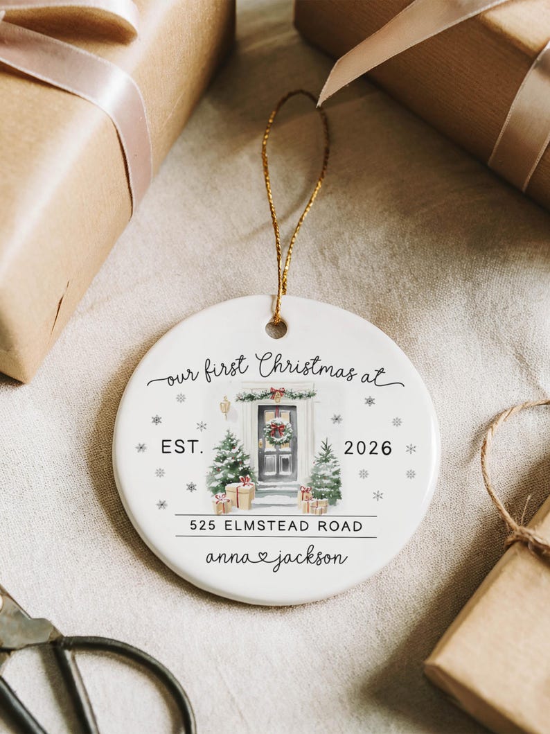 May include: A round white ceramic ornament featuring a watercolor illustration of a front door decorated for Christmas, with the text "our first Christmas at", "EST. 2026", "525 ELMSTEAD ROAD", and "anna & jackson" printed in black, suspended by a gold string.
