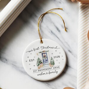 May include: A round white ceramic ornament with a gold hanging string. The design features a watercolor illustration of a blue front door with a wreath, two small Christmas trees, and gifts. Text reads: our first Christmas at EST. 2026 201 SMOKEMONT DRIVE weston family.