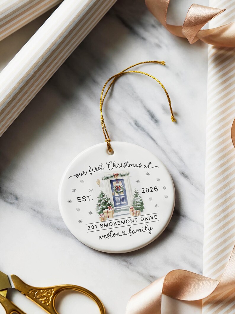 May include: A round white ceramic ornament with a gold hanging string. The design features a watercolor illustration of a blue front door with a wreath, two small Christmas trees, and gifts. Text reads: our first Christmas at EST. 2026 201 SMOKEMONT DRIVE weston family.