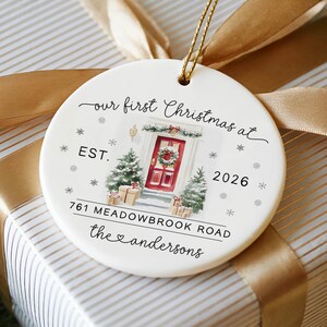 May include: A round white ceramic ornament with a gold hanging string. The design features a watercolor illustration of a red front door with a wreath, two small Christmas trees, and gifts. Text reads: our first Christmas at EST. 2026 761 MEADOWBROOK ROAD the andersons.