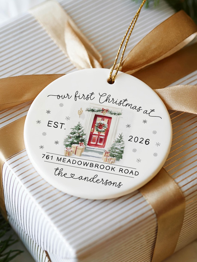 May include: A round white ceramic ornament with a gold hanging string. The design features a watercolor illustration of a red front door with a wreath, two small Christmas trees, and gifts. Text reads: our first Christmas at EST. 2026 761 MEADOWBROOK ROAD the andersons.