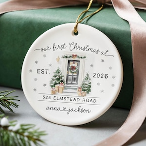 May include: A round white ceramic ornament featuring a watercolor illustration of a front door with Christmas trees, gifts, and snowflakes. Text reads: our first Christmas at, EST. 2026, 525 ELMSTEAD ROAD, and anna heart jackson.