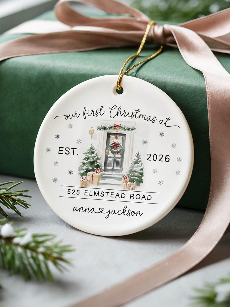 May include: A round white ceramic ornament featuring a watercolor illustration of a front door with Christmas trees, gifts, and snowflakes. Text reads: our first Christmas at, EST. 2026, 525 ELMSTEAD ROAD, and anna heart jackson.