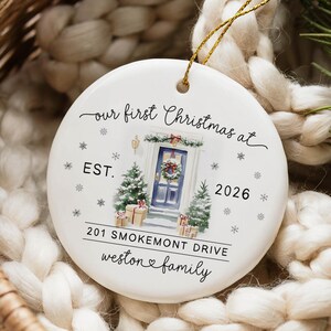 May include: A round white ceramic Christmas ornament featuring a gold hanging string. The design includes a watercolor illustration of a blue front door with a wreath, two small pine trees, and gift boxes. Text reads: our first Christmas at EST. 2026 201 SMOKEMONT DRIVE weston family.