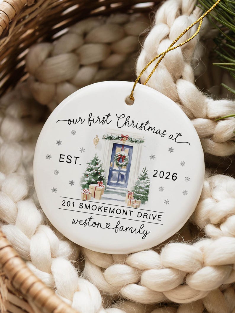 May include: A round white ceramic Christmas ornament featuring a gold hanging string. The design includes a watercolor illustration of a blue front door with a wreath, two small pine trees, and gift boxes. Text reads: our first Christmas at EST. 2026 201 SMOKEMONT DRIVE weston family.