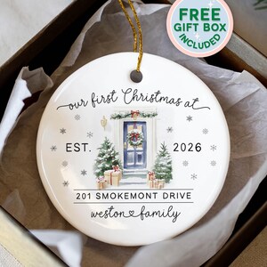 May include: A round white ceramic Christmas ornament with a gold hanging string. The design features a blue front door with a wreath, two snow-covered pine trees, and gift boxes. Text reads: our first Christmas at, EST. 2026, 201 SMOKEMONT DRIVE, weston family.