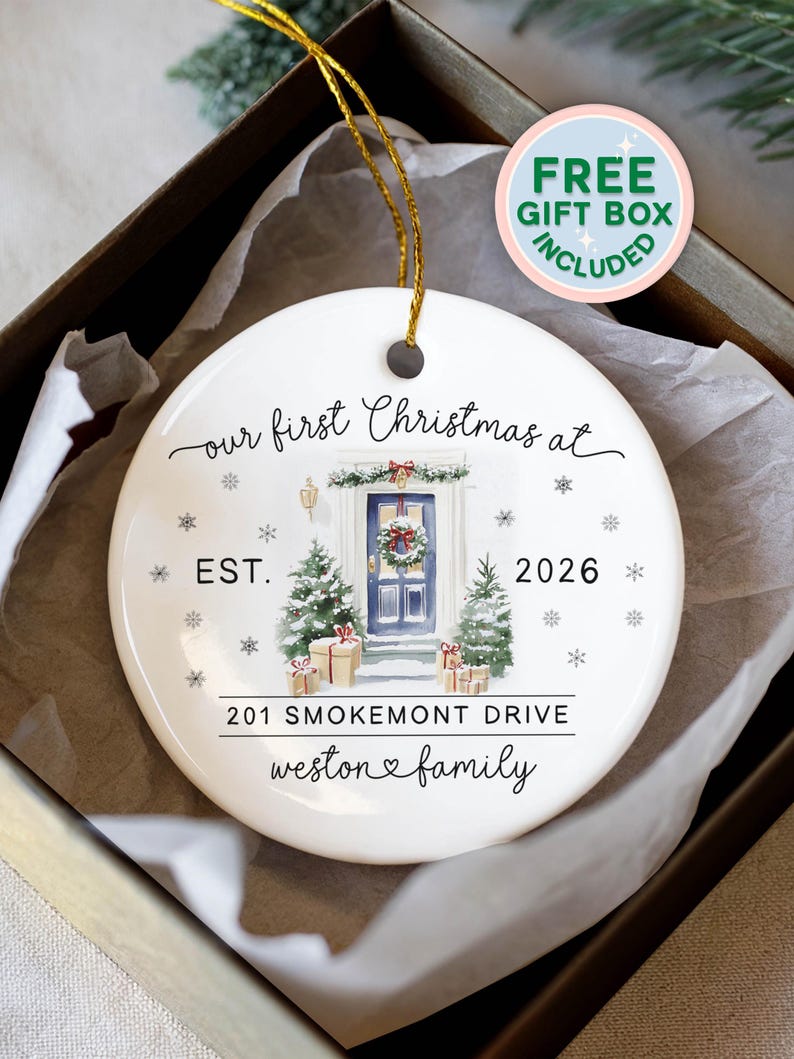 May include: A round white ceramic Christmas ornament with a gold hanging string. The design features a blue front door with a wreath, two snow-covered pine trees, and gift boxes. Text reads: our first Christmas at, EST. 2026, 201 SMOKEMONT DRIVE, weston family.