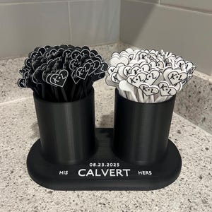 May include: A black and white wedding favor holder with two cylindrical containers. Each container holds heart-shaped toppers with "K+L" and a date. The base reads "HIS CALVERT HERS" and "08.23.2025".