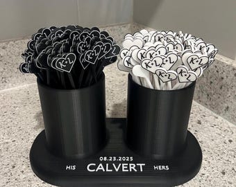 May include: A black and white wedding favour holder with two cylindrical containers. Each container holds heart-shaped toppers with "K+L" and a date. The base reads "HIS CALVERT HERS" and "08.23.2025".