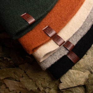 May include: A stack of five folded felted fabric pieces in dark green, burnt orange, cream, light grey, and black. Each piece features a small, rectangular brown leather tag embossed with the text LOVELY CRAFT, stitched onto the folded edge.