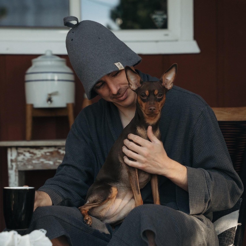 Op de afbeelding: Een grijze sauna hoed met een lus aan de bovenkant. Een persoon in een grijze badjas houdt een kleine bruine en zwarte hond vast. Een zwarte mok staat op een tafel. De sc&egrave;ne speelt zich buiten af.