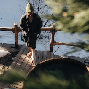 Op de afbeelding: Een persoon gekleed in een donkergrijze badjas, groene hoed en sandalen staat op een houten dek naast een houten hot tub. Het dek heeft een houten reling. Sneeuw en kale bomen zijn zichtbaar op de achtergrond.