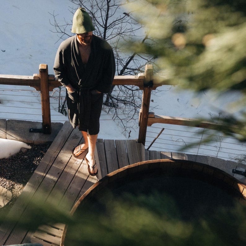 Op de afbeelding: Een persoon gekleed in een donkergrijze badjas, groene hoed en sandalen staat op een houten dek naast een houten hot tub. Het dek heeft een houten reling. Sneeuw en kale bomen zijn zichtbaar op de achtergrond.