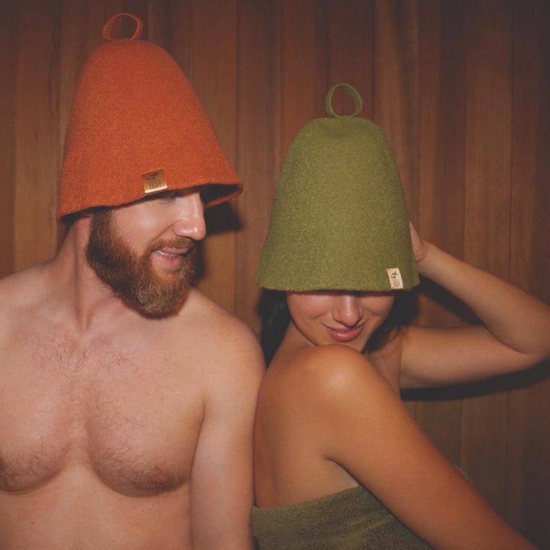 Op de afbeelding: Twee vilten saunahoeden, &eacute;&eacute;n oranje en &eacute;&eacute;n groen, met een kleine lus aan de bovenkant. De hoeden hebben een conische vorm en worden door twee personen gedragen. De achtergrond is een houten sauna.