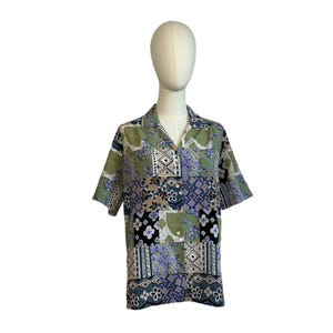May include: A short-sleeved button-down shirt featuring a patchwork design with various geometric and floral patterns in shades of olive green, lavender, black, and white. The shirt has a classic collar and a front button closure with light buttons.