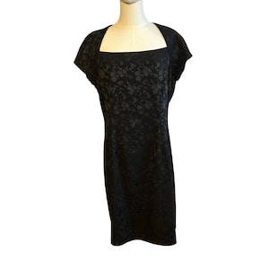 May include: A black knee-length dress with a square neckline and short sleeves. The dress has a subtle floral pattern throughout. The dress is displayed on a mannequin against a white background.