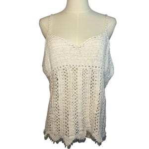 May include: Off-white crochet top with a V-neck and spaghetti straps. The top features a textured, open-weave design with a scalloped hemline. The garment is displayed on a mannequin.