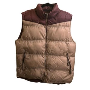 May include: A two-tone puffer vest in tan and burgundy. The vest has a high collar, a full-length zipper, and two zippered pockets. The vest is hanging on a black hanger against a white background.