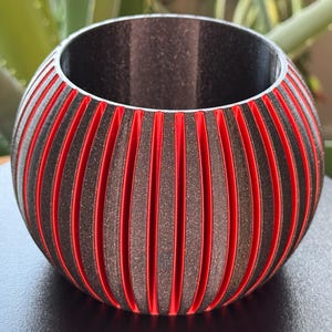 May include: A spherical, decorative container with a ribbed design. The container features alternating vertical stripes of dark grey and bright red. The interior is dark, and the container sits on a dark surface.