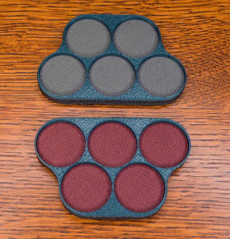 以下が含まれることがあります： 2つのティール色の6穴メイクパレット。1つのパレットには6つの灰色の円、もう1つには6つのマルーン色の円が入っています。パレットは木製の表面にあります。