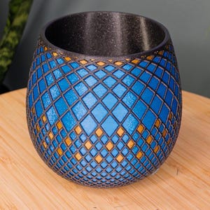 May include: A blue and gold decorative vase with a geometric diamond pattern. The vase has a textured black interior and sits on a wooden surface. The vase is approximately 5 inches tall.