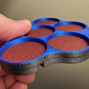 以下が含まれることがあります： 青と黒の3Dプリントされたアイシャドウパレット。5つの円形のくぼみがあり、赤茶色のアイシャドウが詰められています。パレットは手で持たれており、製品のデザインと配色を示しています。
