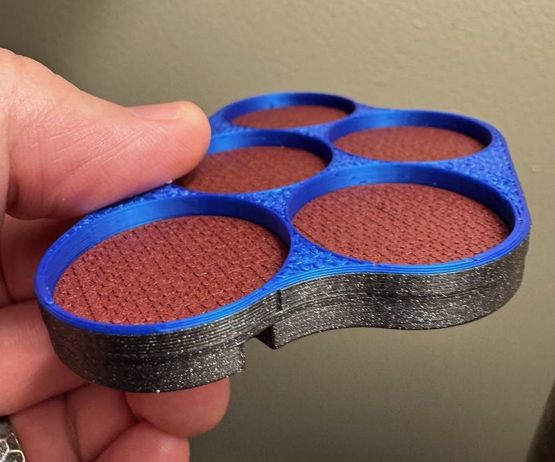 以下が含まれることがあります： 青と黒の3Dプリントされたアイシャドウパレット。5つの円形のくぼみがあり、赤茶色のアイシャドウが詰められています。パレットは手で持たれており、製品のデザインと配色を示しています。