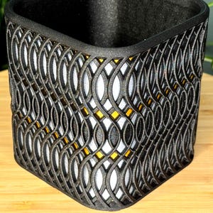 Op de afbeelding: Een zwarte, decoratieve container met een herhalend, verweven patroon. Het ontwerp heeft een reeks onderling verbonden, ovale vormen, waardoor een roosterachtig uiterlijk ontstaat. De binnenkant is wit, met kleine, geeloranje accenten zichtbaar door het patroon.