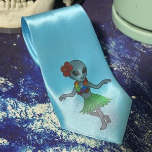 May include: Light blue satin necktie featuring a cartoon alien wearing a flower lei and grass skirt. The alien has a red flower in its hair. The tie is a solid light blue color.