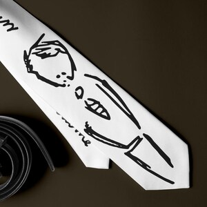 May include: A white tie with a black sketch of a person and handwritten text. The tie is paired with black leather shoes and a rolled-up black belt with a silver buckle. The items are arranged on a dark brown surface.