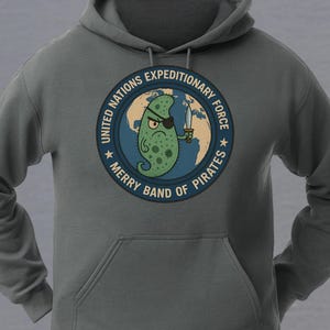 May include: A charcoal gray hooded sweatshirt featuring a circular graphic on the chest. The design shows a green, one-eyed pirate character holding a sword in front of a globe, with the text UNITED NATIONS EXPEDITIONARY FORCE MERRY BAND OF PIRATES.