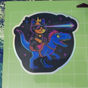 May include: A sticker featuring a cat wearing sunglasses and a crown, riding a blue dinosaur. The cat is shooting a rainbow laser beam. The sticker has a holographic border and a black background with stars.