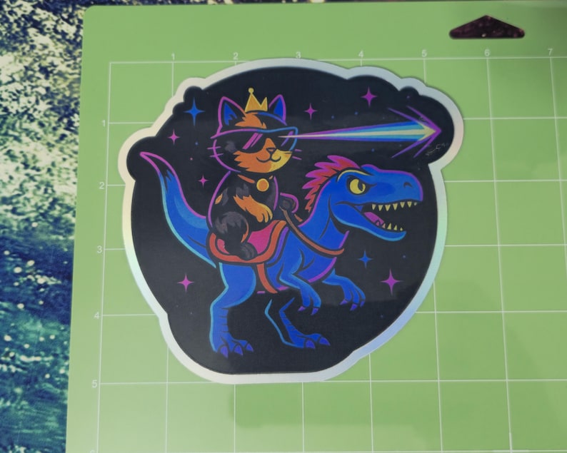 May include: A sticker featuring a cat wearing sunglasses and a crown, riding a blue dinosaur. The cat is shooting a rainbow laser beam. The sticker has a holographic border and a black background with stars.