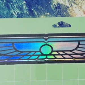 May include: A holographic sticker in the shape of stylized wings, with a central circular design. The wings feature a gradient of blues and greens, with a black outline. The sticker is on a light green cutting mat.