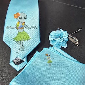 May include: A light blue tie, pocket square, and lapel flower set. The tie and pocket square feature a cartoon alien in a hula skirt. The lapel flower is a solid light blue. The tie has a label that reads "Silk Blend Hand Made Dry Clean Only."