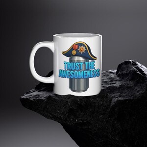 以下が含まれることがあります： 白のセラミックマグカップ。青と金の二角帽をかぶった缶の漫画デザイン。テキスト「TRUST THE AWESOMENESS」は青と白で表示されます。マグカップには、大きくて湾曲したハンドルが付いています。