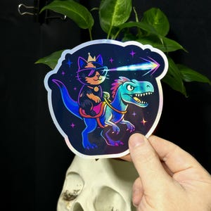 May include: A holographic sticker featuring a cartoon cat wearing sunglasses and a crown, riding a blue dinosaur. The cat is shooting a laser beam. The sticker has a dark blue background with stars.