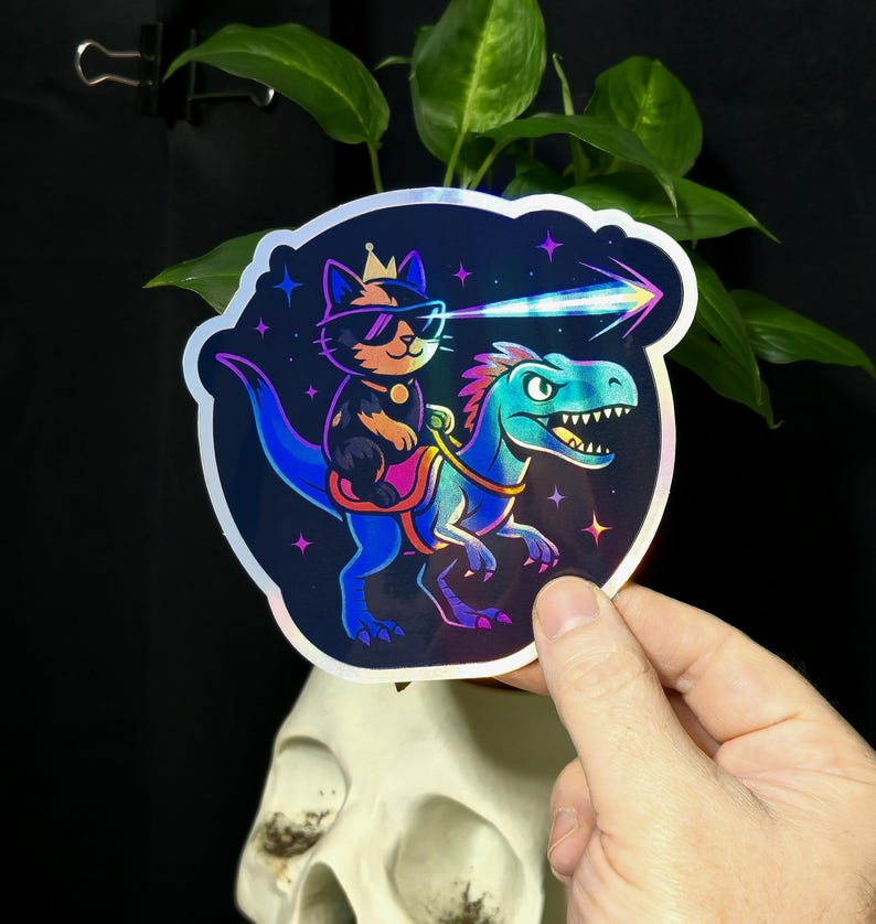 May include: A holographic sticker featuring a cartoon cat wearing sunglasses and a crown, riding a blue dinosaur. The cat is shooting a laser beam. The sticker has a dark blue background with stars.