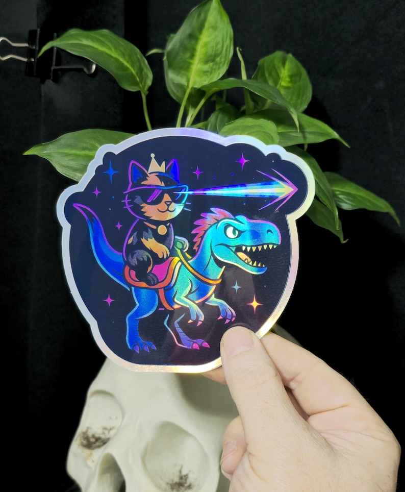 May include: Holographic sticker featuring a cat wearing sunglasses and a crown, riding a dinosaur. The cat is shooting a laser beam. The sticker has a colorful, iridescent finish with a star and space theme.