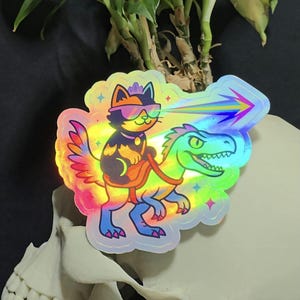 May include: Holographic sticker featuring a cat wearing sunglasses riding a dinosaur. The cat is black and orange, and the dinosaur is green and blue. A rainbow beam shoots from the cat's sunglasses. The sticker has a colorful, iridescent border.