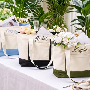 May include: Three canvas tote bags with exterior pockets, each featuring a personalized name embroidered in script. The bags have cream-colored upper sections and contrasting lower panels in light blue, black, and olive green.